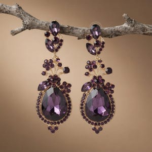 May include: A pair of long, dangling earrings featuring large, pear-shaped purple crystals surrounded by smaller round purple stones set in gold-toned metal. The design includes floral-inspired clusters and teardrop accents hanging from a top crystal.