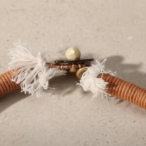 May include: Close-up of a tan rope necklace with white tassel ends. The necklace features a gold-colored clasp with a small, off-white bead. The rope is tightly wound, creating a textured appearance.