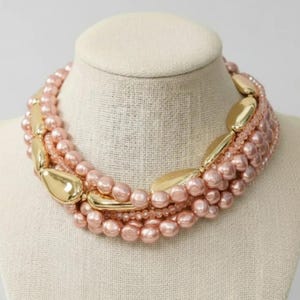 May include: A layered necklace featuring blush-colored faux pearls and gold-toned metal accents. The necklace is displayed on a mannequin bust, showcasing its multi-strand design and elegant aesthetic. The necklace is a statement piece.
