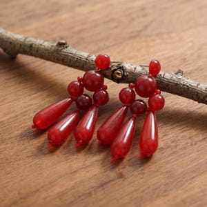 May include: A pair of dangling earrings featuring multiple translucent red beads in varying sizes and shapes, including round spheres and elongated teardrop forms, connected by thin metal components to create a layered, multi-strand design.
