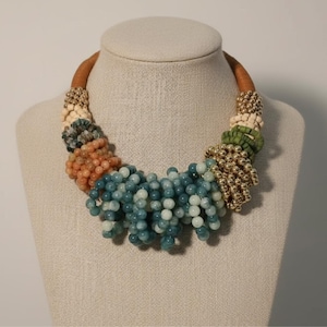 Puede incluir: Un collar llamativo con un collar de cuero marrón. El collar presenta grupos de cuentas azul-verdosas, melocotón, crema y verdes, junto con cuentas de metal dorado. Las cuentas están dispuestas en un diseño en cascada.