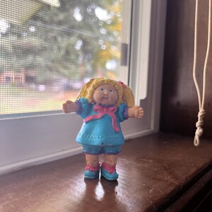 May include: A small plastic figurine of a Cabbage Patch Kid doll. The doll is wearing a blue dress with pink accents and has blonde hair in pigtails.