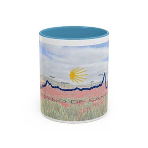 May include: White ceramic mug with a blue interior and a scenic design. The design features a field of red flowers, a blue line graph, and the text "CAMINO DE SANTIAGO". The mug also includes the names of cities.