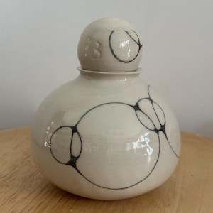 Może przedstawiać: Kremowy ceramiczny słoik o okrągłym korpusie i małej, kulistej pokrywce. Słoik jest ozdobiony czarnymi, abstrakcyjnymi rysunkami liniowymi. Na pokrywce wytłoczona jest liczba "3".