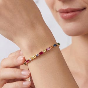 May include: A gold bracelet with a row of colorful rectangular gemstones. The gemstones are in shades of red, pink, yellow, blue, and green. The bracelet is on a wrist.