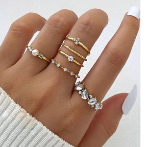 May include: A hand wearing a set of five gold rings with clear and pearl stones. The rings are stacked on the middle finger. The background is white. The text "ILYADAGEMS" is visible in the image.