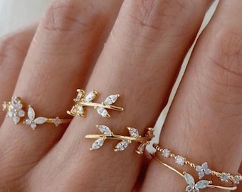 May include: A set of four gold rings with delicate leaf and butterfly designs adorned with clear cubic zirconia stones. The rings are stacked on a finger, showcasing the intricate details of each piece.