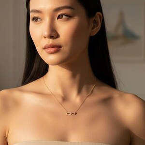 May include: A delicate gold-toned chain necklace featuring a small, horizontal rectangular pendant. The necklace rests against the collarbone, paired with a strapless beige garment. The metal has a polished finish that reflects light.