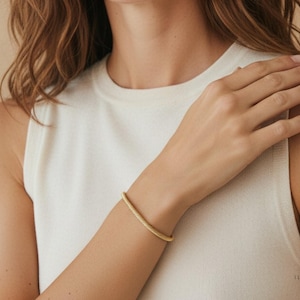May include: A gold-colored bracelet with a twisted rope design. The bracelet is worn on the wrist. The background is a neutral beige color. The person is wearing a white sleeveless top.