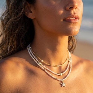 Puede incluir: Un conjunto de collares en capas con tres hilos. El hilo superior es dorado, el hilo del medio tiene cuentas blancas y el hilo inferior tiene cuentas blancas y un colgante de estrella de mar. La marca "ILYADAGEMS" es visible.