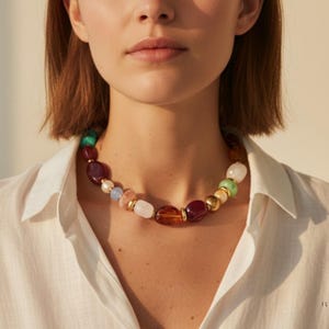 May include: A statement necklace featuring a variety of colourful beads, including red, green, amber, white, and blue. The beads are interspersed with gold-coloured spacers. The necklace is worn against a white collared shirt.