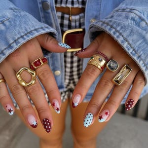 May include: A collection of eight gold-toned rings worn on fingers, featuring various designs including smooth organic shapes, a red rectangular inlay, a wide textured band with embedded stones, a square grey stone, and a long rectangular clear stone.