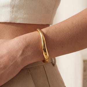 May include: A gold-colored, rounded bangle bracelet. The bracelet is smooth and shiny, reflecting light. The jewelry is worn on a person's arm, with a neutral-toned top and pants in the background. The bracelet appears to be a solid piece of jewelry.