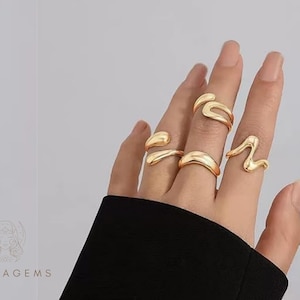 May include: A hand wearing several gold-colored rings with unique, abstract designs. The rings have smooth, flowing shapes, including curved and wave-like forms. The rings are on a hand with long nails, and the background is a light gray.