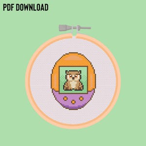 Peut inclure: Un motif de point de croix représentant un Tamagotchi violet et orange avec un hibou brun sur l'écran. Le motif est dans un cercle de broderie.