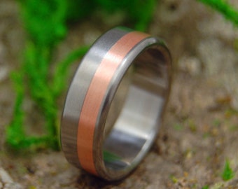 May include: A silver ring with a copper band around the center. The ring is on a brown, textured surface.