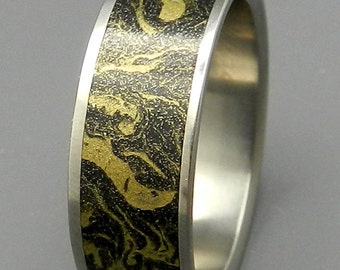 Puede incluir: Un anillo de titanio negro y dorado con un patrón de mármol. El anillo está pulido y tiene un acabado liso.