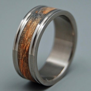 Puede incluir: Un anillo de titanio plateado con una incrustación de madera marrón. El anillo tiene un acabado pulido y un borde biselado.