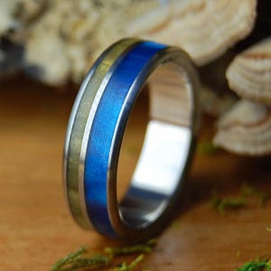 May include: A close-up of a silver ring with a blue and gold inlay. The ring has a polished finish and a modern design. The blue inlay is a vibrant color, and the gold inlay has a textured appearance. The ring is set against a blurred background.