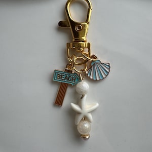 May include: Gold-tone keychain with beach-themed charms. Includes a sign that reads "BEACH", a light blue seashell, a white starfish, and dangling pearls. The keychain is attached to a gold clasp.
