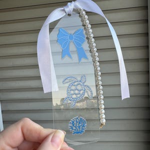 May include: Clear acrylic bookmark featuring a light blue bow, sea turtle, and coral design. A white ribbon and a strand of faux pearls are attached to the top. The bookmark is held up by a hand.