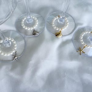 May include: Four wine glasses with pearl beaded charms, each with a different sea-themed pendant: a starfish, a seashell, and a scallop shell. The charms are silver and gold. The glasses are on a white surface.