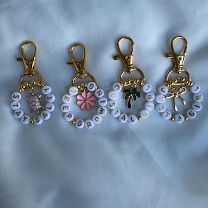 May include: Four keychains with gold-colored clasps and white beaded rings. Each ring has a different word or phrase spelled out in gold letters. Charms include a turtle, a pink flower, a palm tree, and a bow.