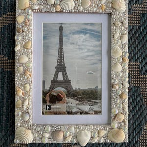 May include: A rectangular picture frame decorated with seashells and iridescent beads. The frame surrounds a photo of the Eiffel Tower. The frame is designed to hold a 4x6 or 5x7 inch photo.