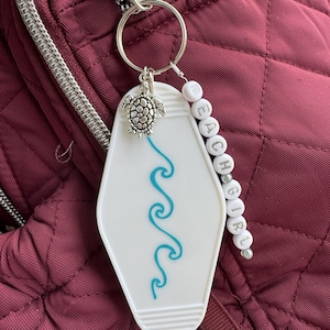 May include: A white keychain with a blue wave design and the words "BEACH GIRL" spelled out in white beads. A silver turtle charm hangs from the keychain, which is attached to a silver keyring. The keychain is set against a burgundy backpack.