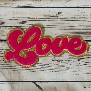 May include: Pink felt "Love" sign with gold glitter trim. The sign is in a retro, cursive font.
