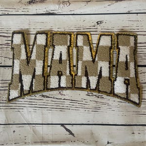 Può includere: Un cartello "MAMA" a scacchi dorati e bianchi con un contorno nero. Il cartello è fatto di tessuto e ha una texture leggermente in rilievo.