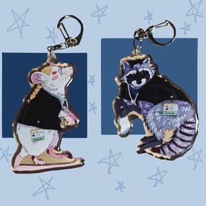Op de afbeelding: Twee acryl sleutelhangers met geïllustreerde dieren in medische kleding. De ene is een witte rat met een blonde vlecht, bril en zwart jasje met een groen 'Doctor' ID-kaartje. De andere is een wasbeer met een stethoscoop en een 'Doctor' ID-kaartje.