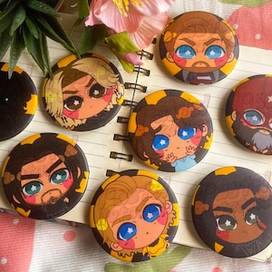 Può includere: Una collezione di bottoni rotondi con personaggi in stile cartone animato, con bordi neri e gialli. Ogni bottone presenta un personaggio diverso con grandi occhi e dettagli colorati. I bottoni sono disposti su un quaderno, con uno sfondo floreale e a motivi di fragole.