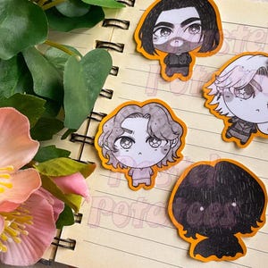 May include: Five anime-style character stickers with orange borders are arranged on an open notebook. The stickers feature various hairstyles and outfits. A potted plant with pink flowers and green leaves is in the foreground.