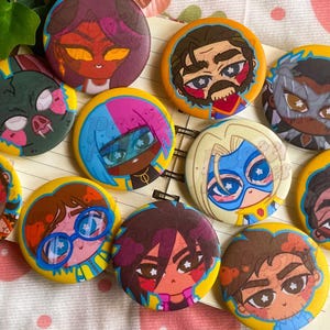 Puede incluir: Una colección de botones redondos y coloridos con varios diseños de personajes de dibujos animados. Los botones tienen bordes amarillos y representan caras con diferentes expresiones y peinados. El fondo es una tela clara con un patrón de fresas.