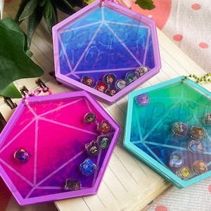 Puede incluir: Tres llaveros coloridos de forma geométrica. Cada llavero tiene un centro transparente lleno de pequeños elementos decorativos. Los llaveros son en tonos rosa, azul y verde, con un diseño geométrico. El fondo incluye un cuaderno y una superficie con estampado de fresas.