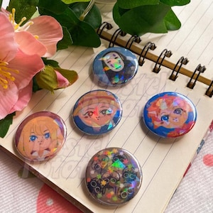 Może przedstawiać: Kolekcja sześciu okrągłych przypinek z postaciami w stylu anime. Przypinki mają różne schematy kolorów i efekty holograficzne. Przypinki są ułożone na notatniku w spiralę, z różowymi kwiatami i zielenią w tle.