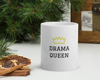 Op de afbeelding: Witte keramische mok met de woorden "DRAMA QUEEN" in zwarte letters, bekroond met een gele kroongrafiek. De mok staat op een wit oppervlak met een gedroogde sinaasappelschijf en kaneelstokjes.