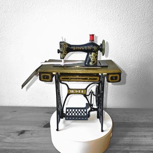 May include: A miniature Singer sewing machine, black with gold detailing, is displayed on a gold-coloured table. The machine has a red thread spool and rests on a white base. The word "SINGER" is visible on the machine's body.