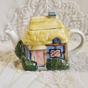Peut inclure: Théière en céramique en forme de cottage. Le toit est jaune, les murs sont bleus et bruns, et la porte est rose. Des buissons verts encadrent le cottage. Elle a un bec et une anse blancs. Objet décoratif charmant.
