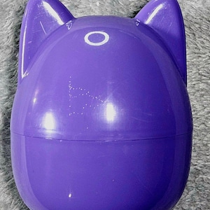 Puede incluir: Un recipiente morado con forma de cabeza de gato, con dos orejas verticales y una abertura circular. El recipiente es de color morado liso y brillante y está sobre una superficie gris texturizada.