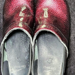May include: A pair of burgundy clogs with a textured, glossy finish. The clogs have a rounded toe and a black sole with the brand name "Dansko" visible. The interior is black with a leather insole.