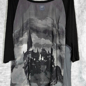 May include: A black and grey raglan-style shirt featuring a graphic print of a castle silhouette against a cloudy night sky. The shirt has solid black sleeves and a scoop neckline with an HP logo printed on the inner collar area of the garment.