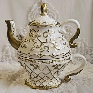 May include: White ceramic teapot with gold accents. The teapot features a decorative gold pattern and a matching lid with a gold finial. The handle and spout are also trimmed in gold. The teapot is designed to hold a single cup.