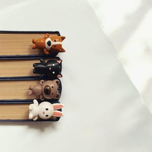 Op de afbeelding: Vier kleurrijke dier-vormige bladwijzers zijn bevestigd aan een stapel boeken. De bladwijzers zijn een bruine hond, een zwarte kat, een bruine beer en een wit konijn.