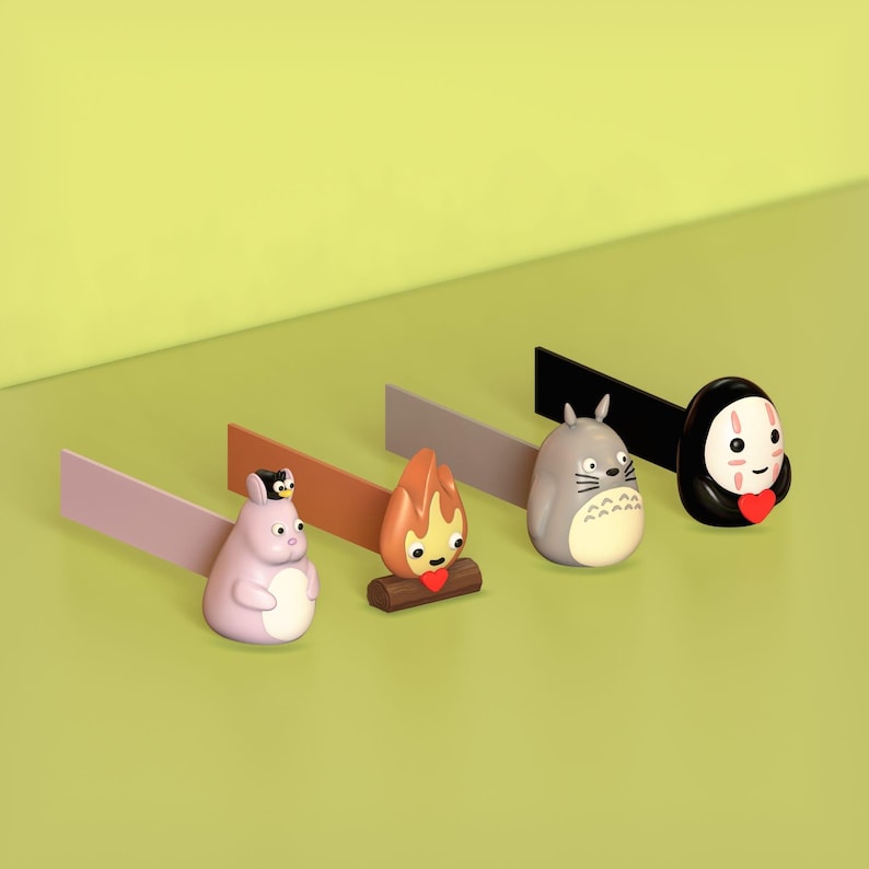 May include: Five colorful character figurines with a small, rectangular sign behind each. The characters are a purple Totoro, a brown and orange campfire, a gray Totoro, a black No Face, and a black and white penguin.