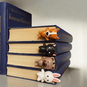 Op de afbeelding: Vier kleurrijke dier-vormige bladwijzers zijn geplaatst op een stapel blauwe hardcover boeken. De bladwijzers zijn een bruine en witte hond, een zwarte kat, een bruine beer en een wit konijn.