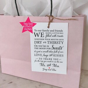 May include: A pink gift bag with a white tag attached. The tag has black text that reads "To our family and friends WE made a little bag filled with treats WHETHER YOUR MOUTH GETS DRY or THIRSTY OR YOU'RE IN THE MOOD FOR Sweets it's just a small token full of our LOVE, hugs and KISSES ~ TO THANK YOU ~ for the trip you made to see the new Mr. and Mrs. Greg and Elsa".