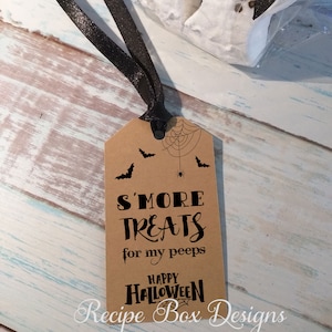May include: A brown paper tag with black text and illustrations for Halloween. The tag reads "S'MORE TREATS for my peeps HAPPY HALLOWEEN" with bat and spiderweb graphics. A black ribbon is attached to the tag.