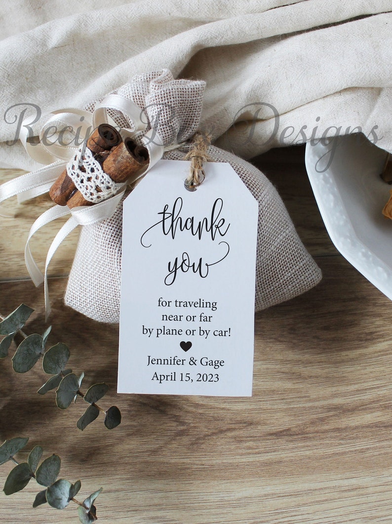 May include: A white thank you tag with black text that reads "thank you for traveling near or far by plane or by car! Jennifer & Gage April 15, 2023". A small black heart is between the names. The tag is tied to a small burlap bag with cinnamon sticks and white lace.